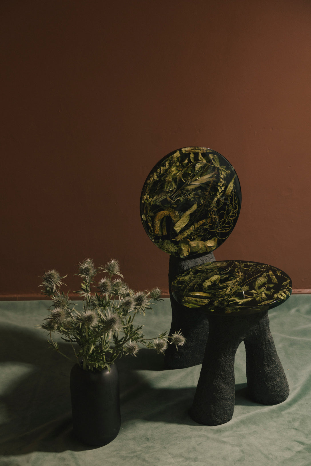 A sculptural chair with a dark green flower resin and black marbled patterned seat and backrest, set against a solid dark brown background on a green fabric surface next to a black vase with dried thistle flowers.