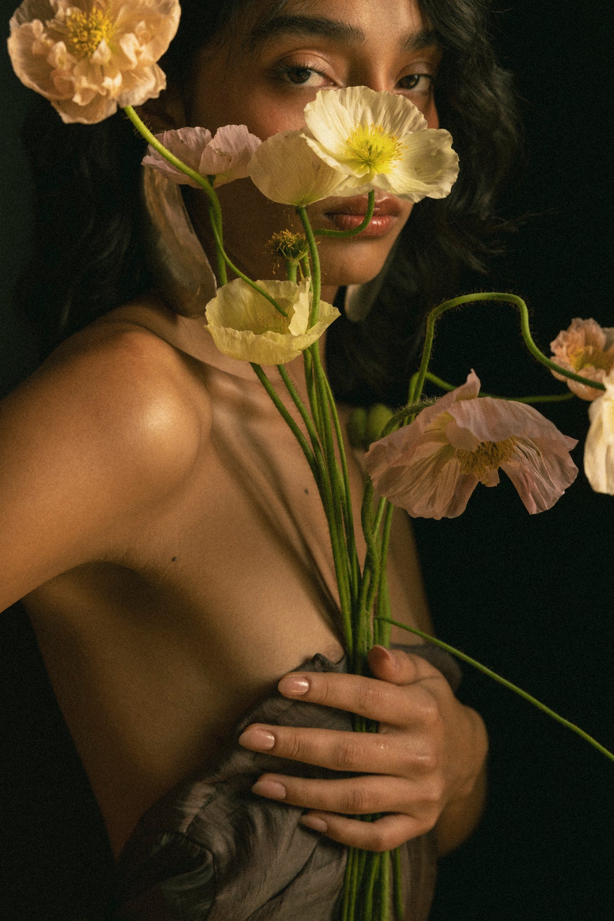 A close-up portrait of a woman holding a bouquet of pale peach and cream flowers against a dark background. Editorial flower shooting